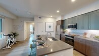North Grove B2F, Unit 5-101, is a thoughtfully designed 2-bedroom, 2-bathroom apartment spanning 1,220 square feet on a single floor in Riverside, CA 92501. The open-concept kitchen and living area serves as the heart of the home, featuring two-tone cabinetry in sage green and warm walnut, stainless steel appliances, subway tile backsplash, and a large center island with bar seating — all set atop light-toned hardwood-style flooring with recessed lighting above. The living space is bathed in natural light from large windows. The primary bedroom (140 sq ft) offers a serene retreat with soft sage walls, a wide window with outdoor views, and access to a well-appointed bathroom with dual vanity, soaking tub, and a walk-in closet with wire shelving. The second bedroom features warm-toned accent walls, abundant natural light, and ample closet space. Both bathrooms are finished with modern fixtures and wood-toned cabinetry. A dedicated laundry room houses a full-size Samsung washer and dryer. The unit also includes an attached garage with an automatic opener, providing secure, private parking. A hallway and entrance area complete this functional, contemporary floor plan.