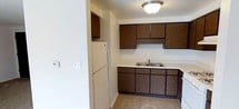 This single-floor residence offers a thoughtfully arranged layout across one bedroom and one bathroom. The main living area features neutral-toned carpeting and clean white walls, creating a bright and open atmosphere. A ceiling fan with a light fixture is centrally positioned overhead, providing both air circulation and ambient lighting to the space.

A partial dividing wall separates the living area from the kitchen, offering a degree of definition between the two spaces while maintaining an open feel. The kitchen is equipped with dark cabinetry, providing a subtle contrast against the lighter surroundings, and includes counter space with a visible sink and backsplash. A refrigerator is also present within the kitchen area.

Adjacent to the living space, a closet with bifold louvered doors offers convenient built-in storage. The neutral color palette and carpeted flooring throughout contribute to a cohesive and understated aesthetic. The layout is efficient and well-organized, making practical use of the available space.

Overall, this residence presents a clean, move-in-ready environment with functional features suited for comfortable everyday living.