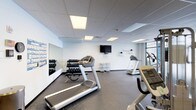 This fitness center offers a comprehensive workout environment designed to accommodate a variety of exercise routines. The space is finished with durable rubber flooring throughout, providing a safe and functional surface for both cardio and strength training activities. A drop ceiling with recessed panel lighting ensures consistent, even illumination across the room, while large windows along one wall invite natural light and offer views of the surrounding exterior.

Cardio equipment is well-represented, with multiple treadmills, an elliptical trainer, and a stationary bike positioned throughout the space. A wall-mounted flat-screen television is installed for entertainment during workouts. A dedicated free weight area features a tiered dumbbell rack accompanied by a full-length mirror, allowing users to monitor their form. A multi-functional cable machine anchors a separate strength training zone, complemented by a stability ball and additional accessories.

The open layout provides ample floor space for stretching or bodyweight exercises. Informational exercise posters and wall charts are displayed for user reference, alongside decorative graphic wall art that adds visual energy to the space. Small utility shelves hold medicine balls and other accessories. A water fountain, bench seating, and a waste receptacle round out the practical amenities, creating a clean and organized fitness environment.