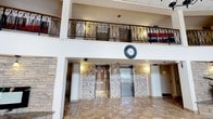 This building lobby makes a strong architectural impression with its soaring double-height atrium design, drawing the eye upward to an ornate wrought-iron balcony railing that runs along the upper level. The scrollwork detailing on the railing adds a classic, refined character to the space, complemented by warm wood accents along the balcony edge. The ground floor is finished with large-format stone-look tile flooring in neutral tones, lending a sense of openness and elegance throughout. A prominent wall clock serves as a functional yet decorative focal point at the center of the lobby, anchoring the space visually. Two elevators are positioned side by side along the rear wall, each framed by decorative mosaic tile surrounds in warm earth tones, with stone veneer columns flanking the elevator bank to add texture and depth. Soft wall-mounted sconces provide warm ambient lighting between the elevator doors, enhancing the inviting atmosphere. The white structural columns and walls provide a clean contrast to the richer decorative elements, while security cameras are discreetly integrated into the design. Overall, the lobby conveys a polished, professional ambiance suited to a multi-story commercial or hospitality building.