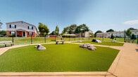 Located in Severn, Maryland, this outdoor dog park offers a thoughtfully designed space for pets and their owners. The park is divided into two separate turf play areas, each covered in lush artificial grass that provides a clean, low-maintenance surface suitable for year-round use. A concrete pathway encircles and separates the two sections, offering easy navigation throughout the space.

Each play area is outfitted with agility-style equipment, including ramp structures, tunnel rings, and decorative rock formations that encourage active play and exploration. Sturdy benches are positioned along the perimeter and throughout the park, giving owners comfortable spots to observe their pets. The entire facility is enclosed by a black metal fence, ensuring a secure environment.

The park is open to the sky, allowing abundant natural light and open views of the surrounding residential community, including mature trees and landscaped grounds that provide a pleasant backdrop. Waste disposal stations are also visible within the space, supporting cleanliness and responsible pet ownership. The layout and amenities reflect a well-planned recreational facility designed to serve both pets and their owners effectively.