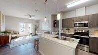 This single-floor residence offers a well-designed open-concept layout that seamlessly connects the kitchen, dining, and living areas. The kitchen is a standout feature, showcasing dark wood-grain cabinetry, light granite countertops, and a large peninsula island with an undermount double sink and elegant pendant lighting overhead. Stainless steel appliances, including a refrigerator, microwave, range, and dishwasher, complete the functional cooking space. Light wood-tone flooring runs throughout the main living areas, enhancing the bright, airy atmosphere.

The living room is bathed in natural light streaming through a set of full-length French doors, which provide outdoor access and contribute to the open feel of the space. A ceiling fan overhead adds comfort, while the neutral wall tones create a versatile backdrop.

The bedrooms are carpeted for warmth and comfort, each featuring windows that allow natural light in and ceiling fans for climate control. The bathroom is well-appointed with wood-tone vanity cabinetry, granite countertops, a large mirror, wood-look flooring, and a shower-tub combination. Throughout the unit, a cohesive neutral palette and modern finishes create a clean, contemporary ambiance.