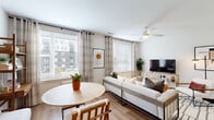 This efficient yet thoughtfully designed 1-bedroom, 1-bathroom residence offers a functional single-floor layout encompassing a living room, kitchen, bedroom, bathroom, hallway, and closet. The open-concept living and dining area is bathed in natural light from multiple large windows dressed with patterned curtains and horizontal blinds, offering views of the surrounding neighborhood. Light wood-tone flooring flows throughout the main living spaces, while a ceiling fan adds comfort overhead. The galley-style kitchen is a standout feature, showcasing charcoal-gray cabinetry extending to the ceiling, sleek white quartz countertops, a subway tile backsplash, stainless steel appliances including a French door refrigerator and range, and recessed lighting. The bedroom provides a comfortable retreat with carpeted flooring, a dedicated closet with built-in shelving and hanging rods, and direct access to the hallway. The full bathroom is finished with light tile flooring, a dark-toned vanity with a white countertop, a tub-shower combination with white tile surround, and recessed lighting, creating a clean and contemporary feel throughout.