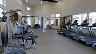 This fitness center is designed to provide a bright, open, and motivating workout environment. The space is defined by its dramatically high ceilings, accented by exposed dark wood beams that add a warm architectural contrast to the otherwise clean, neutral palette of white walls and light flooring. Clerestory windows positioned near the ceiling perimeter allow generous natural light to flood the interior throughout the day, enhancing the airy atmosphere.

The cardio zone is well-appointed with multiple treadmills, ellipticals, and stair climbers arranged in rows, offering ample equipment availability. A wall-mounted flat-screen television provides entertainment for cardio users. The strength training area features a power rack, a dumbbell rack with a full range of free weights, and several selectorized cable and resistance machines suited for a variety of muscle groups.

Large mirrors are strategically placed along the walls, visually expanding the space while allowing users to monitor their form during exercise. The neutral, durable flooring throughout supports both cardio and strength activities. Tall windows and glass-paneled doors provide additional views to the exterior, further reinforcing the open, welcoming ambiance of this well-designed facility.