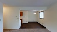 This single-floor space showcases a practical, open-concept layout that connects the living, dining, and kitchen areas seamlessly. The living area features neutral carpet flooring, transitioning to dark hardwood-style flooring throughout the dining and kitchen zones, creating a clear visual distinction between spaces. The walls are finished in a clean, neutral white tone, providing a bright and versatile backdrop throughout.

The dining area is highlighted by a simple chandelier that adds a modest decorative touch, while a window fitted with horizontal blinds allows natural light to filter into the space, enhancing its airy feel. The kitchen, partially visible through an open pass-through wall, features warm-toned wooden cabinetry and includes essential appliances, offering a functional cooking environment.

Dark baseboards provide a grounding contrast against the white walls, adding a subtle design element throughout. The open partition between the kitchen and dining area promotes easy interaction between spaces while maintaining a sense of defined zones. Overall, the space presents a clean, functional layout well-suited for everyday living.