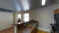This single-floor space is defined by a striking embossed tin ceiling with an intricate repeating pattern that serves as a bold architectural focal point throughout the main living and kitchen area. The metallic ceiling adds historic character and visual depth, contrasting with the neutral cream-toned walls below.

The kitchen is equipped with a wraparound laminate countertop that extends into a peninsula, creating a natural division between the cooking and living areas. The peninsula features a built-in double sink, ample counter space, and dark wood cabinetry below for storage. Upper cabinetry is present near the range area, accompanied by a built-in microwave. The kitchen flows openly into the adjacent living space, enhancing the sense of openness throughout.

Hardwood flooring runs throughout, adding warmth and a classic aesthetic that complements the tin ceiling overhead. Multiple windows fitted with horizontal blinds allow natural light to fill the space, keeping the interior bright and airy. The overall ambiance blends vintage architectural details with functional everyday living elements, creating a characterful and practical environment.