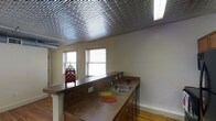 This single-floor space is defined by a striking embossed tin ceiling with an intricate repeating pattern that serves as a bold architectural focal point throughout the main living and kitchen area. The metallic ceiling adds historic character and visual depth, contrasting with the neutral cream-toned walls below.

The kitchen is equipped with a wraparound laminate countertop that extends into a peninsula, creating a natural division between the cooking and living areas. The peninsula features a built-in double sink, ample counter space, and dark wood cabinetry below for storage. Upper cabinetry is present near the range area, accompanied by a built-in microwave. The kitchen flows openly into the adjacent living space, enhancing the sense of openness throughout.

Hardwood flooring runs throughout, adding warmth and a classic aesthetic that complements the tin ceiling overhead. Multiple windows fitted with horizontal blinds allow natural light to fill the space, keeping the interior bright and airy. The overall ambiance blends vintage architectural details with functional everyday living elements, creating a characterful and practical environment.