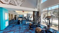 Located at 1401 Montrose Boulevard in Houston's Montrose neighborhood, this single-floor fitness center is a well-appointed exercise facility that combines striking design with comprehensive workout capabilities. The space is defined by its double-height ceilings, which create an open, airy atmosphere, and large floor-to-ceiling windows that flood the interior with natural light and offer views of the surrounding streetscape and mature trees.

A bold pop-art mural spans the upper wall, lending the facility a dynamic, energetic character. Vibrant blue patterned carpeting runs throughout, complementing the modern, urban aesthetic. Recessed cove lighting in a dedicated treadmill zone adds warmth and definition to one section of the space.

The cardio area is well-equipped with multiple treadmills, elliptical machines, a stair climber, and stationary cycling equipment, all positioned to take advantage of the natural light from the expansive windows. A GymRAX functional training wall provides resistance bands, kettlebells, yoga mats, and suspension training accessories for versatile strength and mobility work. The free weights and strength training section features a full dumbbell rack, weight benches, and dual cable/Smith machine systems, supported by mirrored walls that enhance the sense of space. A large-screen display is available for on-demand fitness content, completing this thoroughly equipped facility.