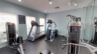 This single-floor fitness room offers a practical and functional space designed to meet essential workout needs. The room is finished with cool blue-gray walls that create a calm, focused atmosphere, complemented by dark rubber flooring that provides durability and traction underfoot. Recessed lighting is installed throughout the ceiling, ensuring the space remains bright and well-lit at all times.

Natural light filters in through several windows fitted with horizontal blinds, adding a welcoming feel to the workout environment. The space is equipped with a range of exercise machines, including a treadmill and an elliptical trainer positioned along the windowed wall for an airy feel during cardio sessions. A multi-purpose cable weight resistance machine is also present, offering users a variety of strength training options. A workout bench is available to complement the strength training equipment.

An informational bulletin board is mounted on the wall, likely displaying fitness guidelines or facility rules. The overall layout is efficient and organized, making good use of the available footprint. This fitness room provides residents or users with a convenient, self-contained space to maintain an active lifestyle without leaving the property.