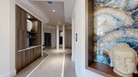 Located at 5400 Broken Sound Blvd NW in Boca Raton, Florida, this upscale wellness spa offers a thoughtfully designed single-floor layout that seamlessly blends modern aesthetics with a serene, restorative atmosphere. The entrance corridor sets an immediate tone of luxury, featuring intricately patterned tile flooring with decorative inlays, warm wood cabinetry, and a striking framed blue-and-gold marbled stone accent panel. A sculptural marble feature wall adorned with dramatic veining serves as a focal point in the main hallway, complemented by an elegant globe chandelier and decorative lanterns that create a refined, tranquil ambiance. A dedicated seating alcove showcases lush floor-to-ceiling greenery walls, bold contemporary artwork, and plush cushioned chairs arranged around a gold accent table, offering guests a relaxing reception area. The central hub of the spa opens into an octagonal-tiled rotunda framed by sleek white columns, providing access to multiple private wellness rooms including a steam room and a cedar-lined dry sauna. Private treatment rooms are finished with wide-plank hardwood flooring, soft ambient wall sconces, vibrant large-scale artwork, and built-in workstations, creating a calm and professional environment for therapeutic services. Every detail of this spa reflects intentional, high-end design craftsmanship.