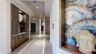 Located at 5400 Broken Sound Blvd NW in Boca Raton, Florida, this upscale wellness spa offers a thoughtfully designed single-floor layout that seamlessly blends modern aesthetics with a serene, restorative atmosphere. The entrance corridor sets an immediate tone of luxury, featuring intricately patterned tile flooring with decorative inlays, warm wood cabinetry, and a striking framed blue-and-gold marbled stone accent panel. A sculptural marble feature wall adorned with dramatic veining serves as a focal point in the main hallway, complemented by an elegant globe chandelier and decorative lanterns that create a refined, tranquil ambiance. A dedicated seating alcove showcases lush floor-to-ceiling greenery walls, bold contemporary artwork, and plush cushioned chairs arranged around a gold accent table, offering guests a relaxing reception area. The central hub of the spa opens into an octagonal-tiled rotunda framed by sleek white columns, providing access to multiple private wellness rooms including a steam room and a cedar-lined dry sauna. Private treatment rooms are finished with wide-plank hardwood flooring, soft ambient wall sconces, vibrant large-scale artwork, and built-in workstations, creating a calm and professional environment for therapeutic services. Every detail of this spa reflects intentional, high-end design craftsmanship.