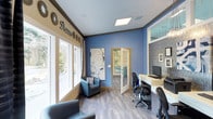 This single-floor space presents a well-designed interior that balances functionality with a welcoming aesthetic. The room is defined by its striking blue accent walls, which are complemented by warm beige trim and dark crown molding, creating a visually dynamic contrast throughout. Decorative wall lettering and circular sunburst accents add a personalized touch to the upper wall area.

Natural light flows generously through large multi-pane windows fitted with patterned valances, offering views of lush outdoor greenery. The windows illuminate the space and enhance its open, airy feel. Smooth light-colored hardwood-style flooring runs throughout, contributing to a clean and modern foundation.

A dedicated workstation area along one wall features a built-in desk surface equipped with a computer monitor, providing an efficient workspace. Multiple ergonomic office chairs are positioned at the desk, accommodating several users simultaneously. A pair of blue upholstered accent chairs offer comfortable seating for visitors or guests.

A set of French doors with frosted glass panels leads to an adjoining room, suggesting additional functional space beyond. An accent wall to the right features textured paneling and framed artwork, adding depth and sophistication. Overall, the space conveys a professional yet approachable atmosphere.