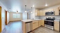This single-floor unit offers a clean, modern aesthetic anchored by light-toned luxury vinyl plank flooring and warm wood-trimmed doorways throughout. The open-concept layout connects the kitchen directly to the living area, creating a spacious and functional flow. The kitchen is well-appointed with light maple cabinetry, light-toned countertops, and a full suite of stainless steel appliances, including a refrigerator, electric range, over-the-range microwave, and dishwasher. A peninsula with a double sink and pendant lighting above serves as a natural divider between the kitchen and living space. The living area benefits from natural light through a window overlooking greenery, complemented by a ceiling fan overhead.

A hallway connects the living and kitchen areas to the private rooms, which include multiple bedrooms, each featuring windows with outdoor views and ceiling fans. One bedroom offers an exceptionally spacious walk-in closet outfitted with wire shelving and hanging rods on multiple walls. The bathroom is designed with accessibility in mind, featuring a large roll-in shower with stainless grab bars. Throughout, the unit maintains a bright, neutral palette that lends a fresh and welcoming ambiance.