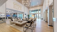 Located at 7950 Park Blvd N in Pinellas Park, Florida, this expansive single-floor community clubroom offers a generously sized, open-plan layout designed for socializing and recreation. The space immediately impresses with its soaring double-height ceilings, shiplap wall accents, and wide-plank hardwood flooring that runs throughout, lending a warm, coastal-modern aesthetic.

The lounge area features a curated arrangement of plush white upholstered chairs and sofas around a substantial wood coffee table, anchored beneath a striking vehicle-themed chandelier. A bold "Good Luck" neon sign adorns the shiplap feature wall, adding a playful, welcoming character to the space.

Moving deeper into the room, a dedicated game zone includes a full-size billiards table and a foosball table, accompanied by industrial-style pendant lighting. A well-appointed kitchen bar area with dark cabinetry, a white quartz countertop, and bar seating provides a functional entertaining hub, while an adjacent dining nook with tufted banquette booths and pendant lighting offers a quieter gathering spot.

Floor-to-ceiling black-framed glass doors and windows flood the interior with natural light and provide direct access to an outdoor patio. Thoughtfully placed artwork and curated furnishings complete the refined yet relaxed atmosphere of this versatile community space.