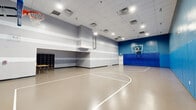 Located at 8601 Chatsfield Way in Hyattsville, Maryland, this spacious indoor athletic facility offers a well-appointed basketball court designed for both competitive and recreational use. The single-floor layout provides an expansive, open playing surface with clearly marked court lines, including painted three-point arcs and lane markings, complemented by a regulation backboard and hoop at each end of the court.

The facility features soaring ceilings with a recessed drop-tile grid housing a robust array of overhead fluorescent and LED lighting fixtures, ensuring consistent, bright illumination across the entire playing surface. The walls are finished in a striking two-tone scheme of blue and grey with bold horizontal striping, adding visual energy to the space. Full-length blue padded wall mats line the lower portions of the court-side walls, prioritizing player safety.

Additional functional elements include a built-in cubby storage unit along one wall, a folding table, and a secondary room adjacent to the main court that can serve as a storage or preparation area. Clearly marked exit doors with safety hardware are positioned throughout the space, and fire safety equipment is visibly installed. The smooth, durable court flooring is built to withstand high-impact athletic activity, making this facility a versatile and well-equipped venue for sports and fitness programming.