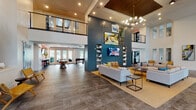 Located at 10000 Palma Linda Way in Orlando, FL, this contemporary community clubhouse delivers an impressive blend of modern design and resort-style amenities. The grand entrance welcomes visitors with floor-to-ceiling glass walls, large-format tile flooring, and abundant natural light that floods the open interior. A dramatic double-height great room serves as the heart of the space, anchored by a bold teal shiplap feature wall adorned with curated artwork and built-in shelving flanking a wall-mounted TV. Generous seating arrangements with neutral sectionals and vibrant accent pillows invite gathering and relaxation beneath a striking mid-century modern chandelier and rich wood-paneled ceiling accents. A staircase with sleek cable railings and warm wood treads leads to an upper mezzanine level overlooking the main lounge. The game room is well-appointed with a foosball table, shuffleboard, a vibrant orange accent wall, bar-height seating, and multiple mounted screens. A separate lounge area features a full kitchen with dark cabinetry, pendant lighting, and an expansive island ideal for entertaining. French doors throughout connect the interior to outdoor areas, including views of a resort-style pool, creating a seamless indoor-outdoor living experience.