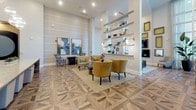 This thoughtfully designed single-floor communal space combines modern aesthetics with functional versatility across several well-appointed areas. The grand entrance corridor sets an elegant tone, featuring soaring ceilings draped with flowing white curtains, gold pendant fixtures, and light stone tile flooring that extends throughout much of the space. Floor-to-ceiling glass at the entry frames outdoor views, filling the hall with natural light.

The main lounge area showcases a striking geometric-patterned hardwood floor, mustard yellow accent chairs, illuminated open shelving displaying curated décor, and large abstract artwork adorning the walls. Adjacent seating nooks offer intimate settings with plush velvet and leather furnishings alongside bold, graphic accent tables.

A dedicated dining area features a generously sized dark wood table surrounded by studded leather chairs, bathed in natural light streaming through tall wraparound windows with sheer drapery. Nearby, a gallery wall displaying framed vinyl record album covers serves as a bold artistic focal point.

The workspace area provides a long built-in desk surface backed by textured teal wallcovering and original artwork. A game and bar area rounds out the space, offering high-top seating and billiards cues mounted on the wall, complemented by etched glass panels featuring tree motifs.
