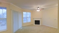 This single-floor space features a well-appointed living room that serves as its central gathering area. The room is finished with wall-to-wall carpeting in a neutral tone, creating a warm and comfortable atmosphere. A white-mantled fireplace with a classic surround serves as a natural focal point, adding both aesthetic appeal and functional warmth to the space.

Crown molding runs along the perimeter of the ceiling, lending a refined architectural detail that elevates the overall look of the room. A ceiling fan with an integrated light fixture is centrally positioned, providing both air circulation and ambient lighting. The walls are painted in a soft, neutral hue that complements the light carpeting and white trim throughout.

A large window fitted with vertical blinds allows generous natural light to fill the room while offering views of the surrounding exterior. A floor vent is visible near the window, indicating a ducted HVAC system. The combination of the fireplace, crown molding, and natural light gives the space a balanced and inviting character suitable for everyday living.