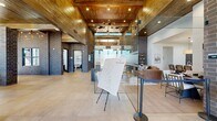 This expansive single-floor residence blends industrial character with modern refinement, offering a thoughtfully designed layout across a generous footprint. The living area makes an immediate impression with soaring double-height ceilings clad in rich wood planking, dramatic exposed brick columns, and large-format tile flooring that anchors the open, airy space. A statement chandelier and recessed lighting add warmth and sophistication to the dramatic volume overhead. Floor-to-ceiling glass partitions define functional zones while maintaining visual openness throughout. A bright office or workspace area is framed by brick columns and expansive windows that flood the room with natural light and exterior views. The bedroom is a serene retreat featuring light wood-style flooring, a ceiling fan, and a well-proportioned window that invites natural light. It is complemented by a dedicated walk-in closet with multi-tier wire shelving for ample storage. Both bathrooms are finished with dark wood cabinetry, granite countertops, large vanity mirrors, and clean white tile tub-shower combinations with herringbone-patterned surrounds. Light wood-style flooring carries throughout these spaces, contributing to a cohesive and polished aesthetic. The hallway connects all areas seamlessly, completing this distinctive and well-appointed residence.