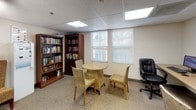 This single-floor community space serves a dual purpose as both a library and a computer room, thoughtfully arranged to support both digital and traditional reading needs. The computer area features a long, continuous workstation desk accommodating multiple desktop computers with monitors, keyboards, and mice, each position equipped with its own adjustable office chair. The setup is well-lit by recessed panel lighting set into a drop ceiling, creating a bright, productive environment.

Transitioning into the library area, tall dark-wood bookcases line the walls and are filled with a broad selection of books spanning various genres. A compact round table surrounded by wicker-style chairs provides an inviting spot for reading or small group conversations. Natural light filters in through windows fitted with horizontal blinds, adding warmth to the neutral-toned, carpeted interior.

Additional amenities include a water dispenser and informational bulletin boards near the entrance, enhancing the room's practical utility. The overall ambiance is calm and functional, with warm neutral wall colors and carpet flooring contributing to a comfortable, welcoming atmosphere suited for focused work, leisure reading, or quiet socializing.