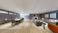 The Mill Lounge is a thoughtfully designed amenity space that seamlessly blends comfort, functionality, and contemporary style across a single, expansive floor. Wide-plank light wood flooring runs throughout, creating a warm and cohesive foundation that unifies each distinct zone.

The main lounge area is anchored by a classic white fireplace flanked by a wall-mounted television, with generous seating arrangements including plush grey sofas, accent chairs in rich cognac leather, and curated artwork displayed along the deep grey walls. Recessed lighting and elegantly detailed ceiling molding contribute to the refined atmosphere.

Adjacent to the lounge, a formal dining area accommodates a large group with a sleek rectangular table and upholstered seating, ideal for gatherings and events. A separate sitting nook features floor-to-ceiling glass doors opening directly to an outdoor terrace, flooding the space with natural light and providing access to fresh air and views of the surrounding landscape.

The well-appointed kitchen includes a large island with seating, open shelving, stainless steel appliances, and a dedicated coffee station, offering everything needed for casual entertaining. A secondary kitchenette with a sink, dishwasher, and open shelving provides additional convenience.

Completing the amenity offering is a fully equipped fitness center with cardio machines, strength training equipment, full-length mirrors, and exterior-facing windows that keep the space bright and energizing.