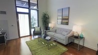 This contemporary two-story space offers a thoughtfully designed interior that balances style and functionality. The living area immediately impresses with its soaring double-height ceilings, creating an airy, open atmosphere. Rich hardwood flooring runs throughout, adding warmth and a sense of continuity to the space. Expansive floor-to-ceiling windows with sheer curtains allow generous natural light to pour in, seamlessly connecting the interior to the lush greenery visible outside. The living room is well-appointed, featuring a plush light grey sofa accented with patterned throw pillows, a sleek glass and metal coffee table, and a vibrant green textured area rug that anchors the seating arrangement. A classic armchair and a tall indoor plant add layers of texture and life to the room. Contemporary wall art adorns the neutral-toned walls, complementing the clean, modern aesthetic. Stylish table lamps provide warm ambient lighting, enhancing the overall inviting atmosphere. The two-floor layout offers distinct living zones, providing both open communal areas and more private spaces above. Throughout, the design strikes a refined balance between modern minimalism and comfortable livability.
