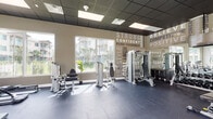 This fitness center is a thoughtfully designed, single-floor facility built to support a comprehensive workout experience. The space is defined by its open layout, dark rubber flooring, and a sleek drop ceiling with recessed panel lighting that provides consistent, even illumination throughout.

One of the most striking design features is the large-scale motivational typography mural that spans an entire wall, displaying words such as "Strong," "Confident," "Believe," "Positive," and "Push Yourself," creating an energizing atmosphere for users. Large windows line multiple walls, flooding the space with natural light and offering views of the surrounding outdoor amenities, including a pool area and landscaped grounds.

The cardio zone is well-appointed with multiple treadmills, an elliptical trainer, and a stationary bike, several of which are positioned to face the outdoor views. A wall-mounted television is available for entertainment during cardio sessions. The strength training area includes a multi-function cable machine, a comprehensive dumbbell rack with a full range of free weights, adjustable benches, and various selectorized resistance machines targeting multiple muscle groups. Additional functional training equipment, including stability balls and balance tools, rounds out the offerings. Glass double doors provide easy access to the exterior, enhancing the connection between the indoor facility and the outdoor environment.