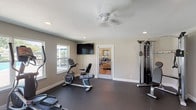 Located at 2301 Windchester St in Pearland, TX, this single-story fitness center is a well-equipped exercise facility designed to support a comprehensive workout routine. The space features durable dark rubber flooring throughout, neutral gray walls, and a textured ceiling with recessed lighting and a ceiling fan, creating a clean and functional environment.

The facility is outfitted with a variety of cardio equipment, including treadmills, an elliptical trainer, and a recumbent stationary bike. Strength training is supported by a multi-station cable machine and weight bench. Additional fitness accessories are neatly organized on a storage rack, including medicine balls, dumbbells, yoga mats, stability balls, and resistance tools, making the space versatile for varied workout styles.

Multiple windows line the walls, allowing ample natural light to fill the room while offering direct views of the outdoor pool area and patio seating, creating an open and inviting atmosphere. A wall-mounted flat-screen television provides entertainment during workouts. An interior door connects the fitness room to an adjacent lounge or common area, and an exterior door with a window panel provides convenient direct access to the outdoor amenity space.