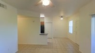 This single-floor space offers a practical and functional layout designed for comfortable everyday living. The main living area is finished with light-toned ceramic tile flooring that runs throughout, providing a clean and cohesive look. A ceiling fan with an integrated light fixture is centrally mounted, ensuring air circulation and ambient lighting across the open space. Natural light filters in through a window on the side wall, brightening the interior and contributing to an airy feel. A semi-open kitchen is accessible from the living area and features a peninsula-style breakfast bar with dark countertops, warm wood-toned upper and lower cabinetry, and a black refrigerator. The kitchen is partially separated from the living space by a half-wall, creating a sense of division while maintaining openness. An additional flush-mount light fixture provides supplemental lighting near the entryway or dining area. A white door off the kitchen area leads to an adjacent room or utility space, adding to the functional layout of the home. The overall palette of soft whites and neutral tones gives the space a clean, understated character suited for a range of uses and personal styles.