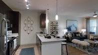 This single-floor space showcases a thoughtfully designed open-concept layout that connects the kitchen and living area in a seamless, functional flow. The kitchen serves as a central feature, anchored by a peninsula with granite countertops that doubles as a breakfast bar with barstool seating — ideal for casual dining or entertaining. A stainless steel sink, dark espresso cabinetry, and built-in appliances contribute to the kitchen's clean, modern aesthetic. Three elegant pendant lights hang above the peninsula, complementing the recessed track lighting overhead to create a well-lit, inviting atmosphere. Light hardwood flooring extends throughout, adding warmth and visual continuity between the spaces. The living area transitions naturally from the kitchen, featuring a neutral color palette accented by decorative elements, including a large framed artwork and a decorative wall-mounted candle holder that adds artistic character. The overall ambiance is contemporary and welcoming, with a balance of functional design and stylish detail that makes the space feel both practical and refined.