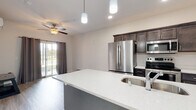 This single-floor unit is designed with a clean, contemporary aesthetic and an efficient, open layout. The kitchen serves as a central feature, showcasing dark espresso cabinetry that extends to the ceiling, light quartz countertops, and a full suite of stainless steel appliances, including a side-by-side refrigerator, electric range, and built-in microwave. A generously sized island with an integrated double sink and pendant lighting above adds both functionality and visual interest. The kitchen flows seamlessly into the living area, which benefits from light wood-look flooring, recessed lighting, and a ceiling fan. A sliding glass door at the far end of the living space allows natural light to filter in and provides outdoor access. The bedroom features plush carpeting, a ceiling fan, a wall-mounted mini-split unit, and a window overlooking the exterior. The bathroom is finished with light tile flooring, a dark vanity with a white countertop, a wall mirror with overhead lighting, and a tub-shower combination. Overall, the unit offers a cohesive, neutral palette with modern fixtures and practical finishes throughout.