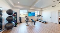 Located at 301 S Calhoun Pl in Star, Idaho, this fitness center offers a thoughtfully designed workout space across a single open floor. The room is finished with light hardwood-style flooring and crisp white walls, creating a clean, energizing atmosphere throughout. Recessed lighting and a ceiling fan provide both illumination and air circulation, while large windows along the perimeter flood the space with natural light and offer views of the surrounding landscape.

The fitness area is well-appointed with a diverse selection of equipment, including a treadmill, stationary bike, adjustable weight bench, a full dumbbell rack, and a medicine ball station with a variety of weighted balls. TRX suspension training straps are mounted to a central structural column, expanding workout versatility. A large wall-mounted flat-screen television adds an entertainment element for exercisers. Full-length mirrors along one wall enhance the sense of openness and support form monitoring during workouts.

Near the entrance, a countertop service area provides convenient access to informational materials and supplies. Glazed door panels with an illuminated exit sign ensure clear and safe egress. An open doorway connects to an adjacent space featuring a kitchen or lounge area, suggesting this fitness center is part of a larger community or commercial facility.