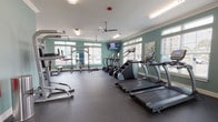 This fitness center at 600 67th Ave N in Myrtle Beach, SC, is a single-level facility designed to support a comprehensive workout experience. The space is finished with soft teal walls, crisp white crown molding, and white baseboards, creating a clean and energizing atmosphere throughout. Rubber flooring covers the main workout areas, providing durability and safety, while the studio section features hardwood-style flooring suited for dynamic movement exercises.

The cardio zone is lined with large windows fitted with horizontal blinds, allowing ample natural light to fill the space. Equipment in this area includes True treadmills, elliptical machines, a stair climber, and a recumbent bike, with a wall-mounted flat-screen television positioned for viewing during workouts. A ceiling fan overhead helps maintain airflow and comfort.

The strength training area houses a Smith machine, a cable/multi-functional rack, a full rack of dumbbells, and adjustable weight benches, offering a thorough selection of resistance training options. Mirrors are strategically placed to assist with form and to visually expand the space.

A separate studio room provides space for group or individual training with spin bikes, stability balls mounted on a wall rack, and open floor space. This room features direct exterior access through a glass-paned door, offering views of an outdoor pool area. A short entry corridor with carpet connects the fitness center to an adjacent amenity space.
