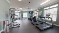This fitness center at 600 67th Ave N in Myrtle Beach, SC, is a single-level facility designed to support a comprehensive workout experience. The space is finished with soft teal walls, crisp white crown molding, and white baseboards, creating a clean and energizing atmosphere throughout. Rubber flooring covers the main workout areas, providing durability and safety, while the studio section features hardwood-style flooring suited for dynamic movement exercises.

The cardio zone is lined with large windows fitted with horizontal blinds, allowing ample natural light to fill the space. Equipment in this area includes True treadmills, elliptical machines, a stair climber, and a recumbent bike, with a wall-mounted flat-screen television positioned for viewing during workouts. A ceiling fan overhead helps maintain airflow and comfort.

The strength training area houses a Smith machine, a cable/multi-functional rack, a full rack of dumbbells, and adjustable weight benches, offering a thorough selection of resistance training options. Mirrors are strategically placed to assist with form and to visually expand the space.

A separate studio room provides space for group or individual training with spin bikes, stability balls mounted on a wall rack, and open floor space. This room features direct exterior access through a glass-paned door, offering views of an outdoor pool area. A short entry corridor with carpet connects the fitness center to an adjacent amenity space.