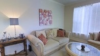 This single-floor space exudes a warm and inviting ambiance, thoughtfully designed with classic elegance and comfortable living in mind. The living area serves as the heart of the space, anchored by a plush, cream-toned sofa adorned with an assortment of decorative pillows in rich earth tones and floral patterns, adding layers of texture and visual interest. A glass-topped oval coffee table complements the seating arrangement, contributing to the room's refined yet approachable aesthetic. Crown molding runs along the ceiling, lending an architectural detail that elevates the overall character of the room. Natural light filters in through a window dressed with horizontal blinds, brightening the neutral wall tones and creating a soft, comfortable glow throughout the space. A small, dark wood side table positioned beside the sofa adds a traditional touch and functional convenience. A vibrant piece of wall art featuring bold circular shapes in shades of coral, pink, and burgundy serves as a focal point, injecting personality and color into the otherwise softly toned interior. The carpeted flooring adds warmth underfoot, completing a space that feels both polished and livable.