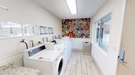 This well-appointed shared laundry facility combines practicality with a lively sense of style across a single floor. The room is outfitted with multiple front-loading washers and top-loading machines arranged in a neat row, offering ample capacity for residents or guests. Light wood-effect tile flooring and crisp white walls create a clean, fresh aesthetic throughout the space, while bright overhead fluorescent lighting ensures excellent visibility at any time of day. A standout feature of the room is a bold, large-scale pop-art mural adorning the back wall, filled with colorful, energetic imagery that adds a dynamic focal point and transforms what might otherwise be a purely utilitarian space into an engaging environment. Large windows along one side of the room allow natural light to filter in, enhancing the open and airy feel. Instructional guides and usage information are posted prominently on the walls for user convenience. A waste receptacle is neatly integrated into the layout, maintaining the room's orderly appearance. The overall ambiance is bright, cheerful, and welcoming, reflecting a thoughtfully designed communal space that balances functionality with visual interest.
