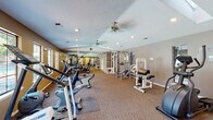 This fitness center is a generously sized, single-level space designed to accommodate a comprehensive range of workout activities. The room features a textured ceiling with multiple skylights that introduce abundant natural light, complemented by recessed lighting and dual ceiling fans that promote air circulation throughout the space. Warm-toned and neutral-gray walls provide a clean, functional backdrop, while wall-to-wall carpeting offers a comfortable surface underfoot.

The facility is equipped with a diverse selection of cardio machines, including treadmills, upright and recumbent stationary bikes, elliptical trainers, and stair climbers. Strength training is well-represented with multi-station cable machines, a leg extension and curl bench, a lat pulldown station, a pull-up and dip tower, and free weight benches. Full-length mirrors are mounted along one wall, enhancing the sense of space while allowing users to monitor their form during exercise.

Large windows line the perimeter walls, offering views of an outdoor pool area and providing additional natural light. A glass-paneled entry door with an illuminated exit sign marks the main access point. The open floor plan allows for comfortable movement between equipment stations, making the space practical and easy to navigate.