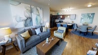 This single-floor residence showcases an open-concept layout that connects the living area and kitchen in one cohesive, flowing space. The living room is anchored by a plush gray sofa accented with decorative pillows in muted yellows and blues, complemented by a sage green accent chair and a solid wood coffee table. A large-scale abstract watercolor artwork adorns the wall, adding a soft, artistic focal point to the room. Warm hardwood-style flooring runs throughout, tying the spaces together seamlessly.

The kitchen is defined by rich dark cabinetry paired with light stone countertops, creating a bold contrast that lends a sophisticated feel. A kitchen island with pendant lighting overhead serves as both a functional workspace and a casual dining area. Black appliances, including a refrigerator and range, complete the well-equipped culinary space. The open sightlines between the kitchen and living area enhance the sense of spaciousness, making the overall layout feel bright, connected, and practical for everyday living.