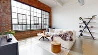 This single-floor industrial loft masterfully blends raw architectural character with contemporary design. Defining the living area are soaring ceilings, polished concrete floors, and dramatic floor-to-ceiling factory-style windows framed in black steel set against original exposed brick walls. The generous living space is bathed in abundant natural light, creating a bright and airy atmosphere with sweeping urban views. Exposed ductwork along the ceiling reinforces the space's authentic industrial aesthetic.

The open-concept kitchen is thoughtfully designed with wood-grain lower cabinetry, clean white countertops, open upper shelving, and stainless steel appliances. A large kitchen island with seating provides an inviting hub for casual dining and entertaining.

The bedroom carries the same industrial character, featuring the signature exposed brick wall and expansive factory windows fitted with blinds for privacy, all anchored by plush carpeted flooring that adds warmth and comfort.

A dedicated hallway connects the living areas to a full bathroom, which features polished concrete floors, a clean white vanity with warm wood cabinetry, and a shower-tub combination. The entryway offers a welcoming transition into the space, complete with a live-edge wood bench and a patterned accent rug.