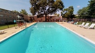 This outdoor pool facility is a standout communal amenity set within a brick-built residential community. At its centerpiece is a generously sized rectangular swimming pool filled with clear, blue-tinted water, designed to accommodate both leisure swimming and active use. Entry ladders are positioned at the pool's edges for easy access, and a lifeguard stand is stationed poolside, underscoring a commitment to safety. A dedicated rescue board and equipment are also mounted nearby, further reinforcing the facility's safety standards.

Surrounding the pool is a broad concrete sun deck that provides ample space for lounging, lined with a variety of colorful deck chairs and seating arrangements. Mature trees rise beyond the fenced perimeter, offering natural shade and a pleasant backdrop to the open-air setting. The area is enclosed by a black iron fence, defining the space while maintaining an open, airy feel.

A shaded seating nook along the brick exterior wall features a picnic-style table with bench seating and a colorful striped umbrella overhead, creating a relaxed spot for socializing. Adirondack chairs nearby add to the casual, welcoming atmosphere. The brick construction throughout the surrounding structures gives the area a classic, enduring character, while the lush tree canopy visible beyond the fence enhances the sense of a green, community-oriented environment.