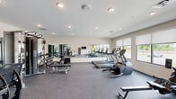 Located at 340 Legion Dr in Lexington, Kentucky, this fitness center offers a thoughtfully designed exercise space that accommodates a wide variety of workout needs. The room features a durable rubber-flooring surface suited for both heavy equipment and dynamic movement, while recessed ceiling lighting ensures consistent, even illumination throughout the space.

Large windows line one wall, flooding the interior with natural light and providing open views of the surrounding outdoor area, creating an energizing atmosphere for workouts. Roller shades offer flexibility in light control when needed.

The cardio zone is well-stocked with treadmills, elliptical trainers, and stationary cycling bikes, all arranged to allow comfortable spacing between users. A full-wall mirror along one side visually expands the space and supports proper form during exercise. The strength training area includes a cable crossover machine, a dumbbell rack with a full weight selection, adjustable benches, and various resistance machines.

A dedicated functional training section features exercise balls, medicine balls, a BOSU balance trainer, and a rowing machine, supporting a diverse range of fitness routines. A wall-mounted television and a water fountain add convenience, while a teal-accented entry door provides a subtle pop of color. Overall, the space is clean, modern, and comprehensively outfitted for an effective fitness experience.