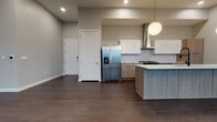 This contemporary high-rise unit is thoughtfully designed with a clean, modern aesthetic throughout. The open-concept kitchen and living area is anchored by a large center island with a light wood finish and white quartz countertops, complemented by two-tone cabinetry combining crisp white upper cabinets with warm wood lower cabinets. A herringbone tile backsplash, stainless steel appliances including a French door refrigerator, a built-in microwave, and a sleek range hood add to the kitchen's refined functionality. Globe pendant lights hang overhead, and rich dark hardwood flooring runs throughout the main living areas. Floor-to-ceiling windows flood the space with natural light and frame striking panoramic views of the city skyline and surrounding mountains. The primary bedroom features plush carpeting, a sliding glass door to a private outdoor space, and direct city views. The primary ensuite bathroom is a spa-like retreat with a freestanding soaking tub, a frameless glass walk-in shower, dual-sink vanity with wood cabinetry, and large-format tile flooring. A secondary bedroom with carpeting, a closet, and city views is served by a well-appointed full bathroom featuring marble-style tile surrounding the tub and shower. A dedicated laundry area with a full-size front-loading washer and dryer completes this polished urban residence.