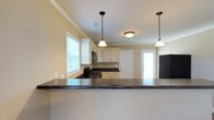 This single-story residence offers 2 bedrooms and 2.5 bathrooms, thoughtfully designed for comfortable everyday living. The kitchen serves as a central highlight, featuring a peninsula with dark granite-style countertops that provide generous prep and serving space. Cream-toned cabinetry with traditional styling lines the walls, complemented by black appliances including a gas range and refrigerator. Two pendant lights hang above the kitchen area, adding focused task lighting alongside a flush-mount ceiling fixture. Crown molding runs along the ceiling perimeter, lending a polished, finished look to the space. A window above the sink area allows natural light to filter in, brightening the kitchen's neutral palette of soft beige walls and white trim. A door adjacent to the kitchen provides convenient exterior access. The open layout between the kitchen and adjoining living areas promotes a connected, functional flow throughout the home. Overall, the space presents a clean and classic aesthetic with practical design elements suited for everyday use.