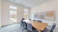 Located at 4020 Chartwell Court in Spring Hill, Tennessee, this professional conference room offers a well-appointed setting designed for productive meetings and collaboration. The space features soaring ceilings with tall transom windows that flood the interior with abundant natural light, providing open views of the surrounding community courtyard. The centerpiece of the room is a substantial natural wood conference table surrounded by upholstered, rolling ergonomic chairs in a cool grey tone, comfortably accommodating groups of up to eight. Patterned, multi-tonal blue-grey carpeting adds texture and warmth underfoot, complementing the clean white walls throughout. A curated triptych of abstract artwork in muted earth and gold tones adorns one wall, lending a refined, contemporary aesthetic to the space. A dark wood credenza provides additional storage and surface space along one wall, accented by a deep navy accent wall detail near the entry. The room is accessed through sleek glass double doors, creating a sense of openness and connectivity to adjacent areas. Recessed lighting supplements the natural light, ensuring a bright and welcoming atmosphere at any time of day. This conference room combines modern design with functional elements, making it an ideal venue for meetings and professional gatherings.