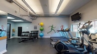 Located at 101 Rantoul St in Beverly, MA, this fitness center is a well-appointed, single-floor facility designed to support a wide range of workout routines. The space features a clean, modern aesthetic with light gray walls accented by vibrant athletic murals depicting running figures, lending the room an energetic and motivating atmosphere. Durable dark flooring runs throughout, providing a practical surface suited for both cardio and strength training activities.

The cardio zone is generously equipped with multiple treadmills, elliptical machines, a water rower, and stationary spin bikes, each positioned near windows with natural light filtering through slatted blinds. Wall-mounted flat-screen televisions are positioned throughout for entertainment during workouts. A dedicated strength area houses a full rack of kettlebells and dumbbells in varying weights, along with stability balls and a medicine ball rack for functional training. TRX suspension straps are mounted to a wall-anchored rig, enabling bodyweight and resistance training exercises.

The facility also includes mirrored walls to support form-checking during exercise, and exposed ductwork along the ceiling adds an industrial character to the space. An AED station, hand sanitizer dispensers, and double-door entry with frosted glass panels reflect a commitment to safety and accessibility. The overall layout is open and well-organized, creating an inviting environment for a comprehensive fitness experience.