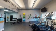 Located at 101 Rantoul St in Beverly, MA, this fitness center is a well-appointed, single-floor facility designed to support a wide range of workout routines. The space features a clean, modern aesthetic with light gray walls accented by vibrant athletic murals depicting running figures, lending the room an energetic and motivating atmosphere. Durable dark flooring runs throughout, providing a practical surface suited for both cardio and strength training activities.

The cardio zone is generously equipped with multiple treadmills, elliptical machines, a water rower, and stationary spin bikes, each positioned near windows with natural light filtering through slatted blinds. Wall-mounted flat-screen televisions are positioned throughout for entertainment during workouts. A dedicated strength area houses a full rack of kettlebells and dumbbells in varying weights, along with stability balls and a medicine ball rack for functional training. TRX suspension straps are mounted to a wall-anchored rig, enabling bodyweight and resistance training exercises.

The facility also includes mirrored walls to support form-checking during exercise, and exposed ductwork along the ceiling adds an industrial character to the space. An AED station, hand sanitizer dispensers, and double-door entry with frosted glass panels reflect a commitment to safety and accessibility. The overall layout is open and well-organized, creating an inviting environment for a comprehensive fitness experience.