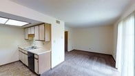 This single-floor space features an open-concept layout that connects the kitchen and living area, creating a cohesive and functional environment. The kitchen is positioned to one side and includes light-toned upper and lower cabinetry with a wood-grain finish, a countertop with a built-in sink, and space for appliances. A half-wall partition separates the kitchen from the adjoining living area, maintaining an open feel while defining each zone.

The living area is generously proportioned, finished with wall-to-wall carpeting in a neutral gray tone that contributes to a quiet, understated atmosphere. Cream-colored walls throughout keep the space feeling bright and clean. A textured popcorn ceiling adds a classic touch, while centrally placed HVAC vents ensure climate control across the unit.

A doorway visible at the far end of the living area leads to additional rooms, suggesting a practical, straightforward floor plan. Baseboards and wall outlets are consistently placed, reflecting attention to utility. Overall, the space presents a clean, no-frills design well-suited for everyday living.