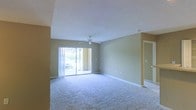 This single-floor space features an open and airy living area finished with wall-to-wall carpeting in a neutral tone, contributing to a clean and comfortable atmosphere. The room is anchored by a ceiling fan with a light fixture, providing both airflow and overhead lighting. A textured popcorn ceiling adds a classic touch to the space.

Natural light flows generously into the room through a large sliding glass door, which offers a view of an outdoor area with greenery and what appears to be a patio or courtyard setting. Vertical blinds accompany the sliding door, allowing for adjustable light control and privacy.

The walls are painted in a warm beige tone, complementing the neutral carpet and creating a cohesive, inviting color palette throughout. White baseboards frame the perimeter of the room, providing a crisp contrast against the flooring and walls. A hallway visible on the right side of the space suggests additional rooms beyond the main living area. Overall, the space offers a functional, well-lit layout with a welcoming ambiance suited for comfortable everyday living.