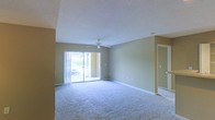 This single-floor space features an open and airy living area finished with wall-to-wall carpeting in a neutral tone, contributing to a clean and comfortable atmosphere. The room is anchored by a ceiling fan with a light fixture, providing both airflow and overhead lighting. A textured popcorn ceiling adds a classic touch to the space.

Natural light flows generously into the room through a large sliding glass door, which offers a view of an outdoor area with greenery and what appears to be a patio or courtyard setting. Vertical blinds accompany the sliding door, allowing for adjustable light control and privacy.

The walls are painted in a warm beige tone, complementing the neutral carpet and creating a cohesive, inviting color palette throughout. White baseboards frame the perimeter of the room, providing a crisp contrast against the flooring and walls. A hallway visible on the right side of the space suggests additional rooms beyond the main living area. Overall, the space offers a functional, well-lit layout with a welcoming ambiance suited for comfortable everyday living.
