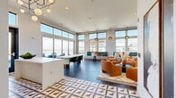 Located at 9305 Montview Blvd in Aurora, Colorado, this thoughtfully designed living room lounge offers a vibrant and inviting atmosphere defined by bold contemporary interiors and an abundance of natural light. Floor-to-ceiling windows wrap the space on multiple sides, framing sweeping outdoor views and flooding the interior with daylight throughout the day.

The living area is anchored by a striking shiplap accent wall featuring a built-in fireplace and wall-mounted television, creating a cozy yet modern focal point. Four plush amber-toned lounge chairs arranged around a dark circular coffee table form an intimate seating cluster, accented by a geometric patterned area rug. A built-in banquette upholstered in teal lines one windowed wall, accompanied by bistro-style side tables and pendant globe lighting overhead.

A set of large folding glass doors opens directly onto a furnished outdoor terrace, seamlessly blending indoor and outdoor living. The entry area features an eye-catching geometric encaustic tile floor, a white waterfall-edge counter, and a kitchenette with teal cabinetry, blush tile backsplash, and a bold chevron-striped accent wall overhead. Dark slate-toned floor tiles ground the main lounge area, while recessed and pendant lighting complete the refined, design-forward aesthetic throughout.